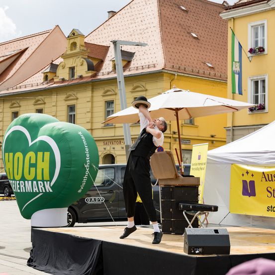 Ein Straßenkünstler steht auf der Bühne und hebt einen Hut mit dem Kinn. Dahinter stehen zwei grüne herzförmige Luftballons. Ein Lieferwagen und Zelte sind geparkt.