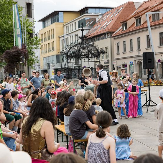 Eine Menschenmenge beobachtet einen Straßenkünstler im Freien. Er hält einen Hut und führt eine Darbietung für das Publikum auf. Dahinter befinden sich Gebäude, Bäume und ein Pavillon.