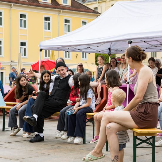 Ein Mann führt eine Show in einem offenen Bereich mit einem Zelt und gelben Bänken auf. Mehrere Kinder und Erwachsene schauen zu. Die Kinder sitzen auf den Bänken, während die Erwachsenen stehen.