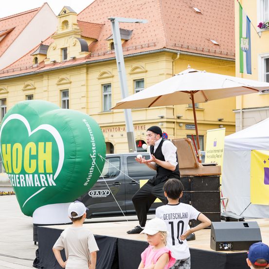 Ein Mann jongliert, während eine Gruppe von Kindern auf einer Bühne zuschaut, mit einem großen grünen Ballon im Hintergrund. Hinter der Bühne steht ein Gebäude und ein Zelt.
