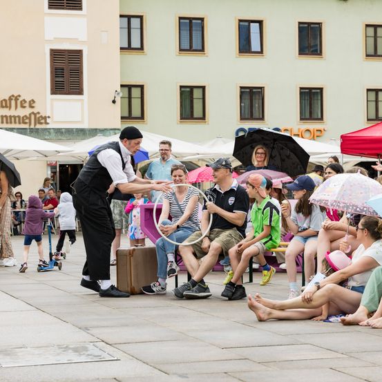 Ein Straßenkünstler unterhält eine Menschenmenge auf einer Freiluft-Plaza. Leute sitzen auf Bänken und schauen zu, wie der Künstler mit einem Hula-Hoop jongliert. Einige halten Regenschirme, andere tragen Hüte. Gebäude mit Schildern säumen den Hintergrund.