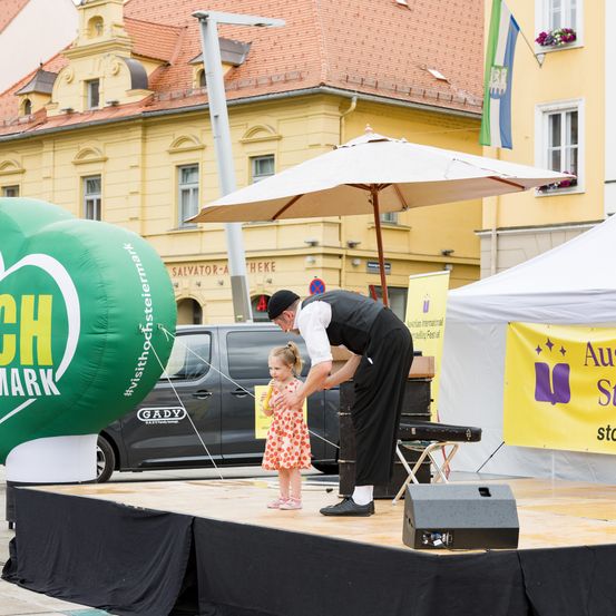 Ein Mann in Schwarz steht mit einem kleinen Mädchen auf der Bühne. Die Bühne ist mit schwarzem Tuch bedeckt, und in der Nähe steht ein Lautsprecher. Im Hintergrund befindet sich ein grüner Ballon, und dahinter steht ein gelbes Gebäude und ein geparkter Van.