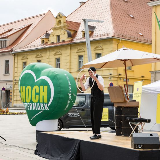 Ein Mann auf der Bühne hält ein Seil und führt eine Zirkusvorführung mit einem herzförmigen Ballon mit der Aufschrift 'HOCH STEIERMARK' durch. Hinter ihm sind ein großer Regenschirm und ein Auto zu sehen.
