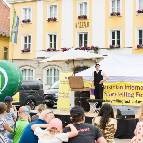 Ein Geschichtenerzähler steht auf der Bühne vor einem Publikum. Die Bühne ist unter einem Schirm, und ein Banner steht Austrian Internal Storytelling Festival.
