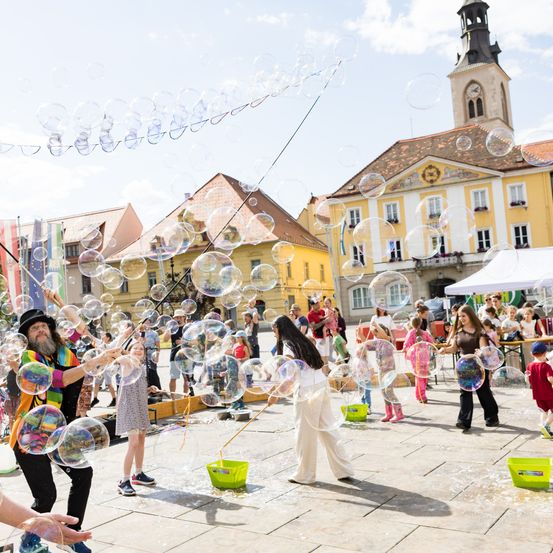 Eine Gruppe von Menschen bläst Seifenblasen auf einem Stadtplatz, mit bunten Gebäuden und einem Turm im Hintergrund. Einige tragen Hüte, und es gibt Seifenwassereimer in der Nähe.