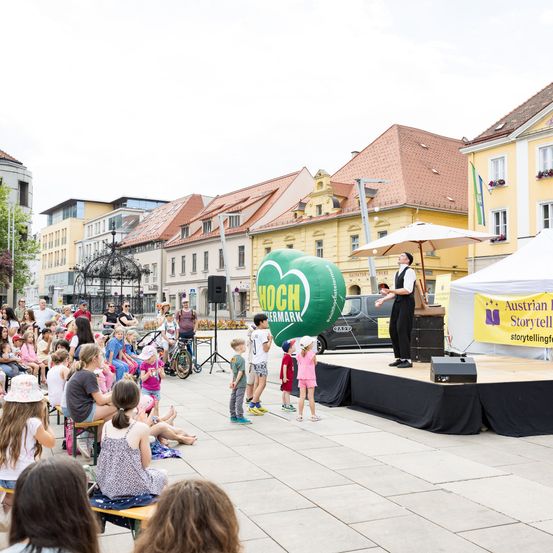 Eine Gruppe von Kindern und Erwachsenen sitzt vor einer Bühne mit einem Künstler. Ein großer grüner Ballon ist über der Bühne. Gebäude sind im Hintergrund.