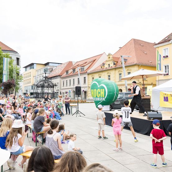 Ein Mann unterhält eine Menge von Kindern auf einem Stadtplatz mit einem großen Luftballon und einem Schirm. Gebäude säumen die Straße, und ein gelbes Auto ist in der Nähe geparkt.
