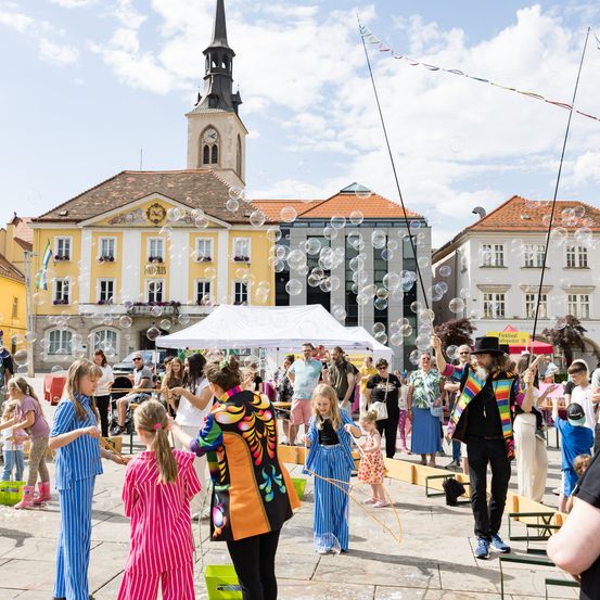 Eine Gruppe von Menschen versammelt sich auf einem Außengelände mit Gebäuden im Hintergrund, einige machen mit Stöcken Blasen, ein Mann ist in einem bunten Outfit und eine Frau in Blau hält einen Stock.