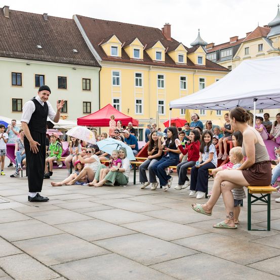 Ein Mann in Schwarz tritt auf einem Platz mit einer Menschenmenge von Kindern und Erwachsenen unter einem weißen Zelt auf. Einige sitzen, andere stehen oder sitzen auf Bänken.