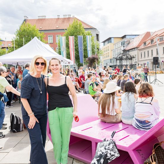 Zwei Frauen stehen neben einer pinkfarbenen Bank in einem überfüllten Außenbereich mit Menschen, Bäumen und Fahnen im Hintergrund.