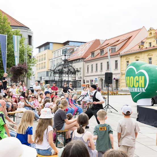 Eine Menschenmenge sieht sich eine Aufführung in einem Außenbereich an. Ein erwachsener Mann tritt auf der Bühne mit einem Mikrofon und einem Lautsprecher auf. Hinter der Bühne steht ein grüner Ballon. Ein paar Leute stehen hinter der Menge. Ein paar Gebäude befinden sich hinter der Menge.
