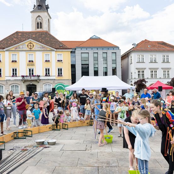 Eine Straßenaufführung findet auf einem Stadtplatz statt. Kinder nehmen mit Hilfe von Erwachsenen an der Aktivität teil. Gebäude und Bäume sind im Hintergrund zu sehen.