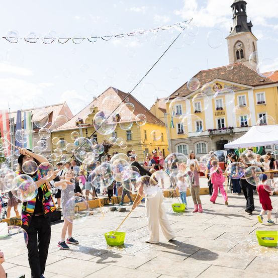 Menschen versammeln sich auf einem offenen Platz mit schwebenden Seifenblasen. Ein Mann trägt ein farbenfrohes Outfit und bläst Blasen. In der Nähe spielen Kinder mit Seifenblasen und grünen Eimern. Im Hintergrund befinden sich Gebäude mit Balkonen und einem Turm.