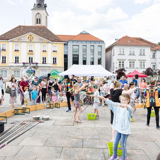 Eine Gruppe von Kindern und Erwachsenen macht Seifenblasen auf einem Stadtplatz. Im Hintergrund befinden sich Gebäude und ein Turm.