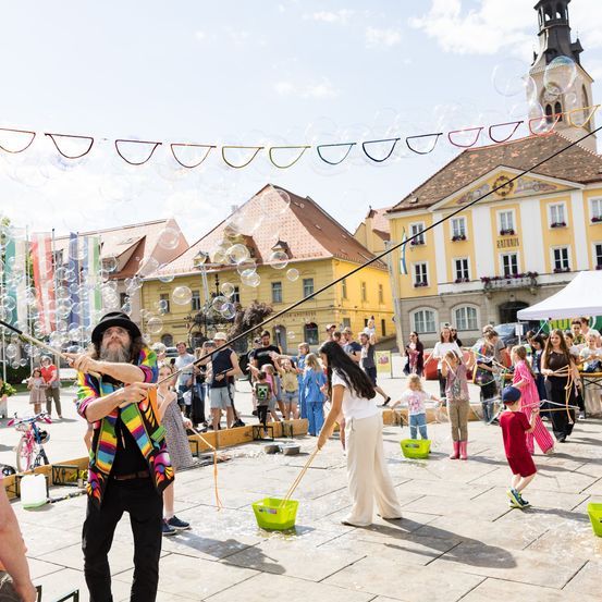 Ein Mann erzeugt in einem öffentlichen Platz Seifenblasen. Viele Kinder und Erwachsene beobachten und spielen mit den Seifenblasen. Im Hintergrund befinden sich Gebäude und Straßenlaternen.