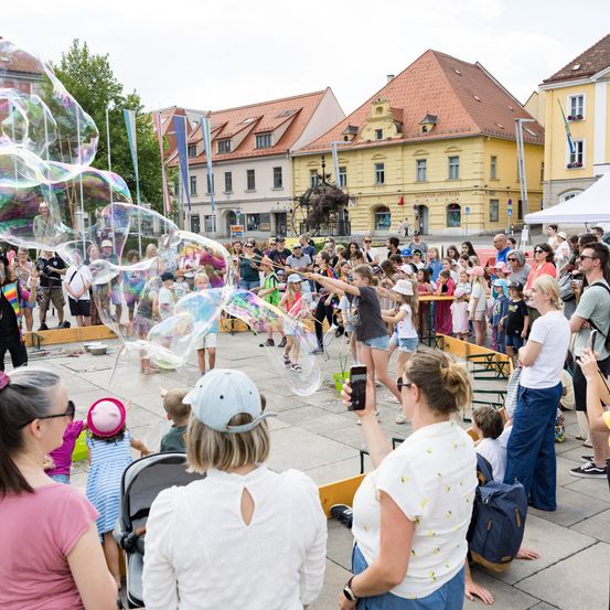 Eine Menschenmenge, einschließlich Kinder, versammelt sich um eine Straßenaufführung mit großen Seifenblasen in einer Freiluftumgebung. Mehrere Personen machen Fotos.