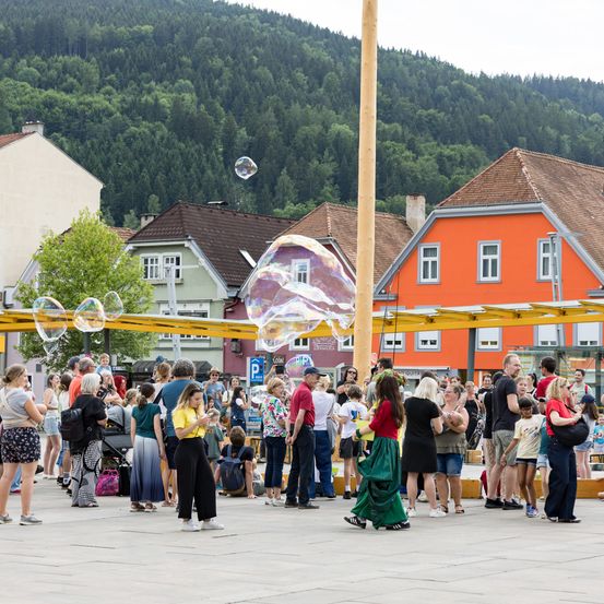 Eine Menschenmenge versammelt sich auf einem offenen Platz in der Stadt. Sie schauen sich riesige Seifenblasen an. Gebäude mit braunen und orangefarbenen Dächern sind im Hintergrund.