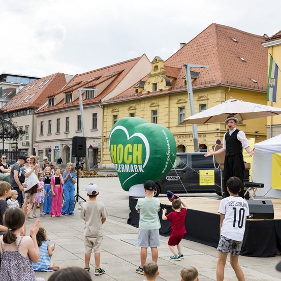 Eine Gruppe von Kindern und Erwachsenen versammelt sich um einen großen grünen Ballon auf einer Stadtstraße. Ein Künstler mit Hut und Hula-Hoop-Reifen tritt auf der Bühne vor ihnen auf.