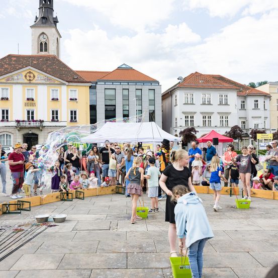 Eine Menschenmenge versammelt sich um eine Seifenblasenmaschine auf einem Stadtplatz. Viele Kinder spielen mit Seifenblasen, während andere zuschauen. Im Hintergrund befinden sich Gebäude und ein Turm.