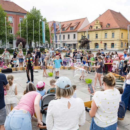 Eine Menschenmenge versammelt sich auf einem Stadtplatz für eine Veranstaltung mit einem Künstler, der Seifenblasen macht. Einige Kinder und Erwachsene schauen zu, während andere an Aktivitäten teilnehmen. Gebäude mit roten Dächern umgeben den Bereich, und Fahnen sind auf Stangen sichtbar.