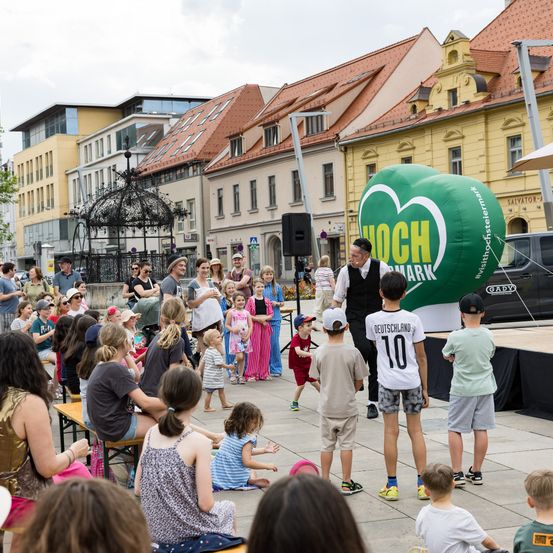 Eine Menschenmenge, darunter Kinder und Erwachsene, hat sich um eine Bühne mit einem grünen Luftballon versammelt. Einige schauen zu, andere gehen auf dem Bürgersteig. Im Hintergrund stehen Gebäude.