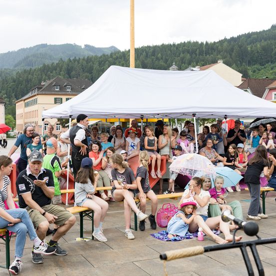 Eine Gruppe von Menschen, darunter Erwachsene und Kinder, versammelt sich unter einem Zelt im Freien. Sie sitzen auf Bänken und einige stehen. Ein Mann steht vorne, möglicherweise in einer Aufführung. Im Hintergrund befinden sich Berge.