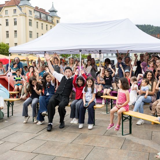 Eine Gruppe von Menschen, einschließlich Kinder und Erwachsene, versammelt sich unter einem weißen Zelt. Einige sitzen auf Bänken, während andere stehen, alle klatschen und jubeln. Ein Mann in der Mitte, gekleidet in einem schwarzen Weste, führt für die Menge auf.