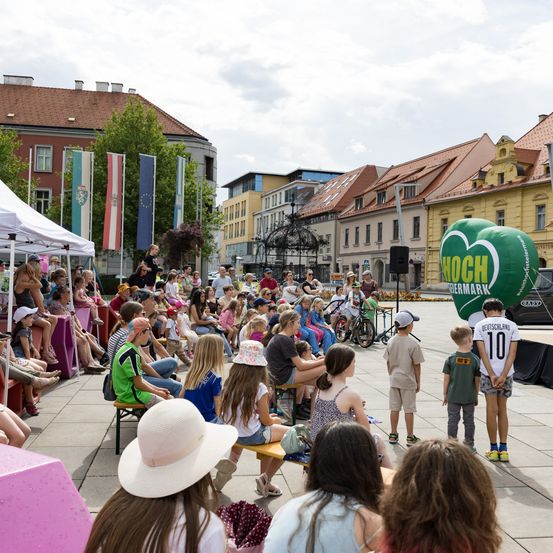 Eine Menschenmenge versammelt sich auf einer Freifläche mit einem großen grünen Luftballon, der Hoch-Tiermann beschriftet ist. Menschen sitzen auf Bänken und einige stehen, um eine Veranstaltung zu beobachten. Im Hintergrund befinden sich Gebäude mit Fahnen.