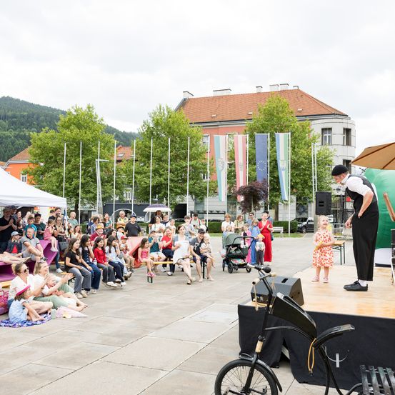 Ein Straßenkünstler unterhält eine Menschenmenge aus Kindern und Erwachsenen auf einer Bühne in einem Park. Flaggen und ein großer Regenschirm stehen hinter dem Künstler.
