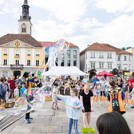 Ein Straßenkünstler erzeugt große Seifenblasen, während eine Menge Kinder und Erwachsene zuschauen. Im Hintergrund befinden sich Gebäude mit roten Dächern und ein Glockenturm.