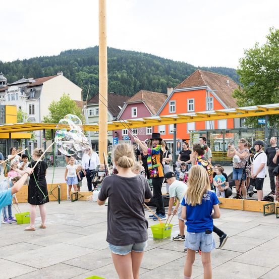 In einem Stadtplatz mit Gebäuden und Bergen im Hintergrund findet eine Straßenaufführung statt. Ein Mann und Kinder machen Seifenblasen.