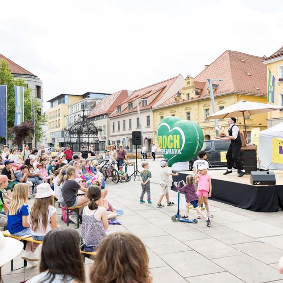 Eine Gruppe von Kindern beobachtet einen Künstler auf einer Bühne mit einem Ballon, während Erwachsene und andere Kinder auf Bänken sitzen und einige herumlaufen. Gebäude mit roten Dächern sind im Hintergrund.