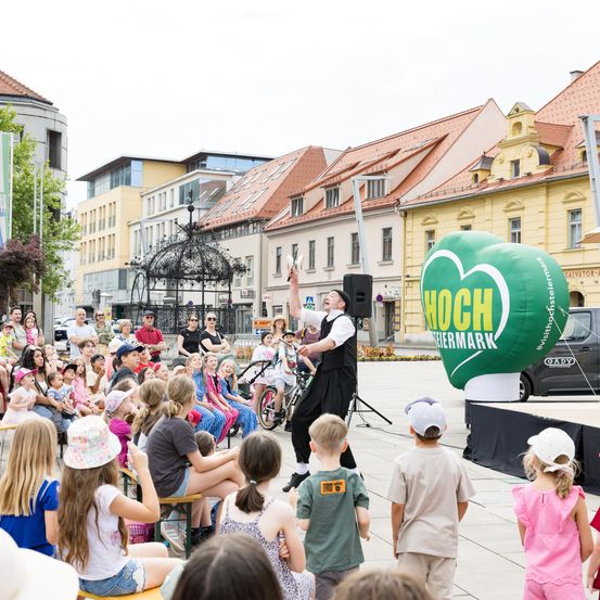 Eine Menschenmenge aus Kindern und Erwachsenen beobachtet einen Künstler auf dem Stadtplatz. Der Künstler hält einen Ball und einen Ballon. Gebäude mit roten Dächern sind im Hintergrund.