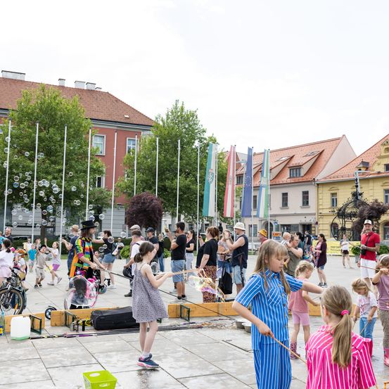 Kinder spielen mit Seifenblasen bei einer Freiluftveranstaltung. Ein Mann in bunter Kleidung steht in der Nähe. Bäume und Gebäude säumen den Hintergrund.