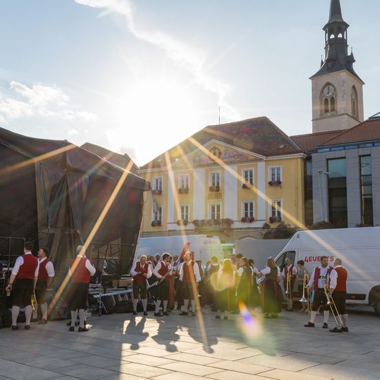 Eine Gruppe von Musikern in traditioneller Kleidung tritt auf einer Bühne vor einem Gebäude auf, mit einem Van und Zelten in der Nähe.