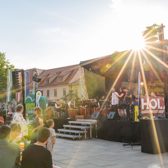 Eine Band tritt auf der Bühne im hellen Sonnenlicht auf. Mehrere Leute schauen von unten zu. Ein Banner mit dem Wort 'HOL' ist auf der rechten Seite sichtbar.