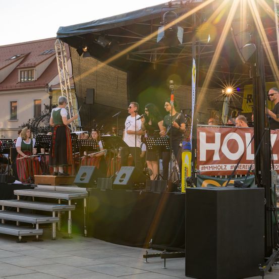 Eine Band spielt auf einer Freiluftbühne mit einem Banner, das HOLY steht. Die Band besteht aus mehreren Musikern, die verschiedene Instrumente spielen.