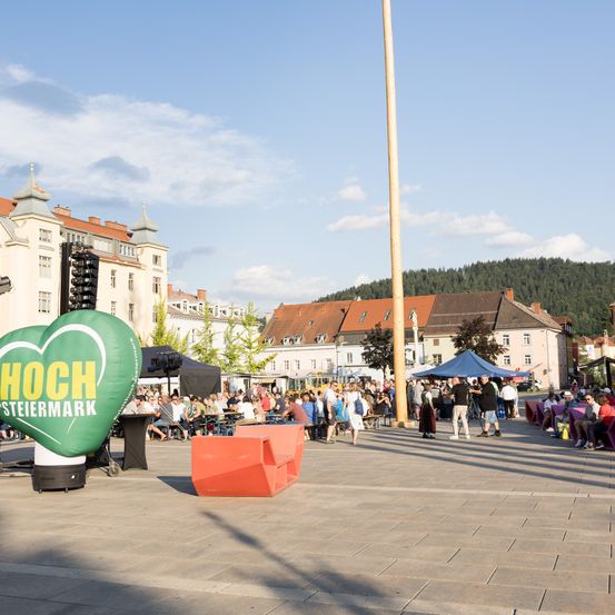 Eine Menschenmenge versammelt sich um ein grünes Herzluftballon für eine Veranstaltung, mit Gebäuden, einem Berg im Hintergrund und einem klaren Himmel.