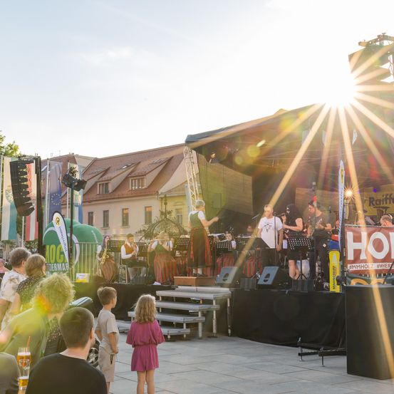 Eine Band spielt auf der Bühne bei einer Outdoor-Veranstaltung, mit Zuschauern und heller Sonneneinstrahlung. Hinter der Band ist ein Gebäude mit Dach und Flaggen zu sehen.