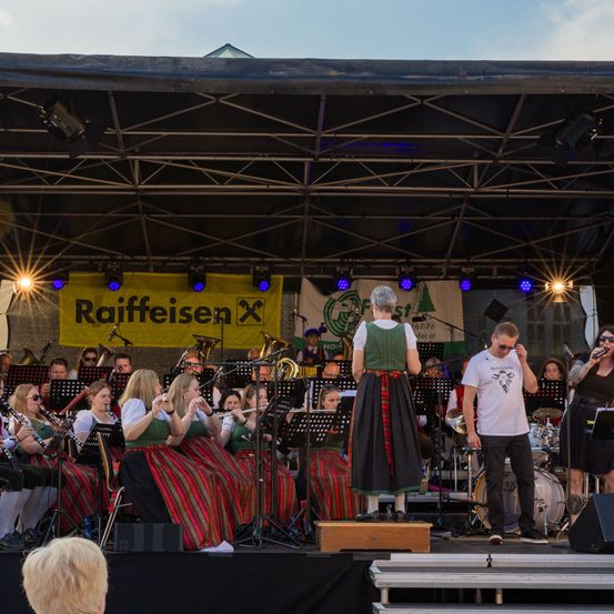 Eine Band spielt auf einer Bühne mit Zeltdach. Sie spielen verschiedene Instrumente. Eine Frau in traditioneller Kleidung steht vorne. Hinter ihnen befindet sich ein Banner mit dem Text Raiffeisen.
