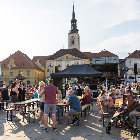 Ein Stadtplatz mit einem Turm im Hintergrund. Ein schwarzes Zelt ist für ein Konzert aufgebaut, viele Menschen sitzen an Bänken und Tischen.