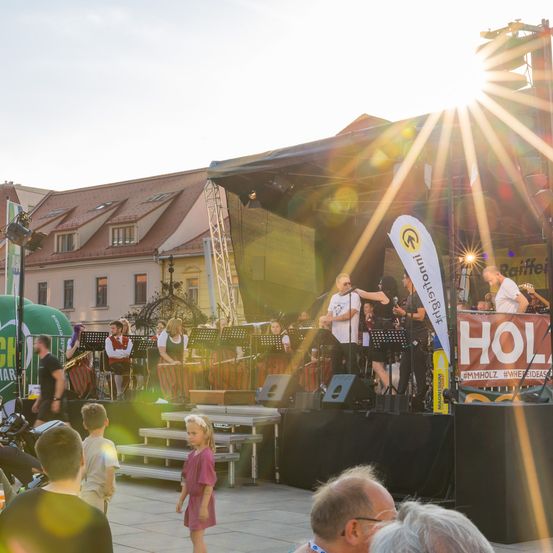 Eine Band spielt auf einer Bühne vor einem Gebäude. Die Sonne scheint hell und wirft Strahlen auf das Publikum. Ein kleines Mädchen steht vor der Bühne und schaut zu.