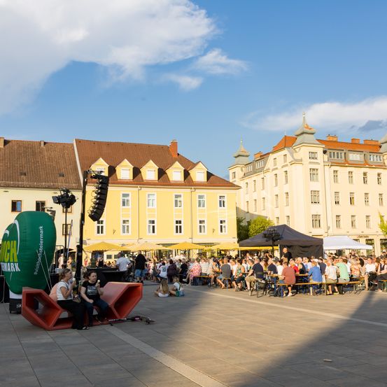 Ein lebendiges Open-Air-Event mit Menschen an Tischen unter Markisen auf einem Stadtplatz, mit Gebäuden im Hintergrund und einem klaren Himmel.