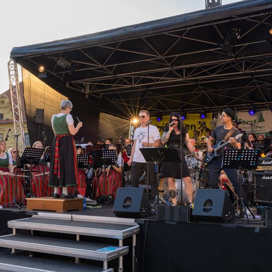 Eine Band tritt auf einer Bühne vor einem großen Publikum auf, mit einem Gitarristen, Sänger und Dirigent, mit Musikinstrumenten und einem schwarzen Hintergrund.