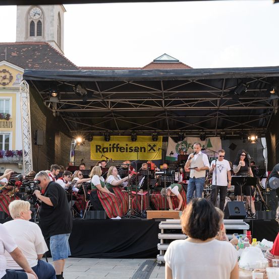 Eine Freiluftveranstaltung zeigt eine Bühne mit einem großen Banner, auf dem Raiffeisen steht. Eine Band spielt mit einem Dirigenten, während das Publikum von verschiedenen Positionen zuschaut. Im Hintergrund ist ein Uhrenturm zu sehen.