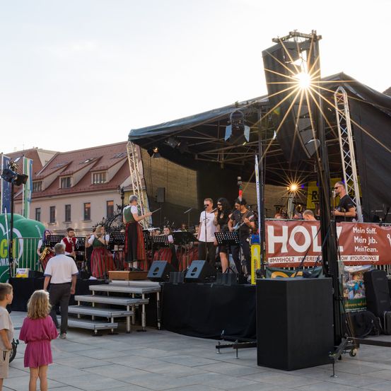 Eine Outdoor-Bühnenaufstellung mit einer Band, die in traditioneller Kleidung auftritt. Die Bühne ist mit einem Zelt abgedeckt, und eine große Menge schaut zu. Scheinwerfer leuchten, und eine riesige Bierflasche ist sichtbar.