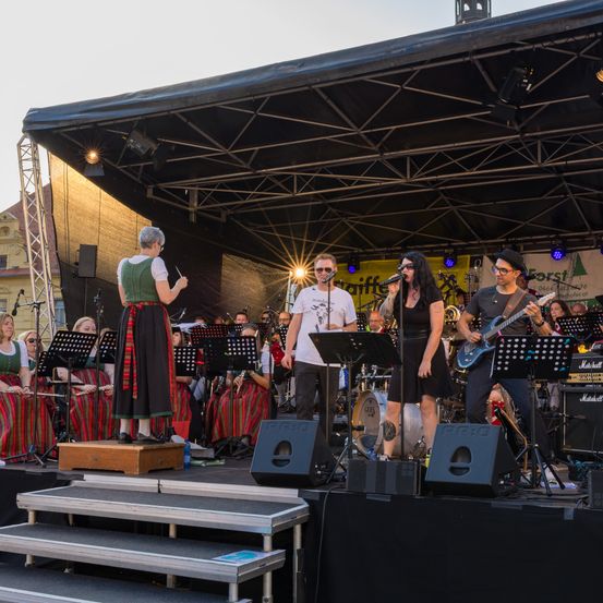 Eine Band tritt auf der Bühne auf, mit einer Sängerin und einem Gitarristen. Dahinter spielen Musiker Blasinstrumente. Scheinwerfer beleuchten die Bühne, und das Publikum schaut zu.