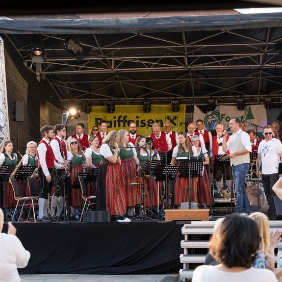 Eine Gruppe Musiker, einige in traditioneller Kleidung, spielt auf der Bühne mit Instrumenten. Scheinwerfer und Notenständer sind sichtbar. Das Publikum sitzt im Vordergrund.