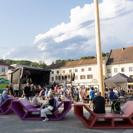 Eine Gruppe von Menschen genießt Outdoor-Sitzplätze bei einem Festival. Lila und rote Bänke sind um den Bereich herum angeordnet, und eine Bühne ist für Aufführungen eingerichtet. Bäume und Gebäude umgeben die Szene.