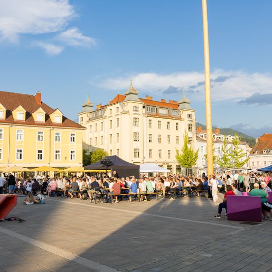 Ein geschäftiger Stadtplatz mit Menschen an Tischen, umgeben von historischen Gebäuden und Bäumen.